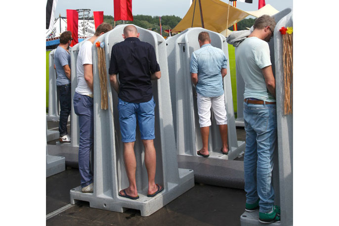 Mehrere Männer stehen in einzelnen, grauen Urinalen im Freien, im Hintergrund sind rote Fahnen und ein Zelt zu sehen.