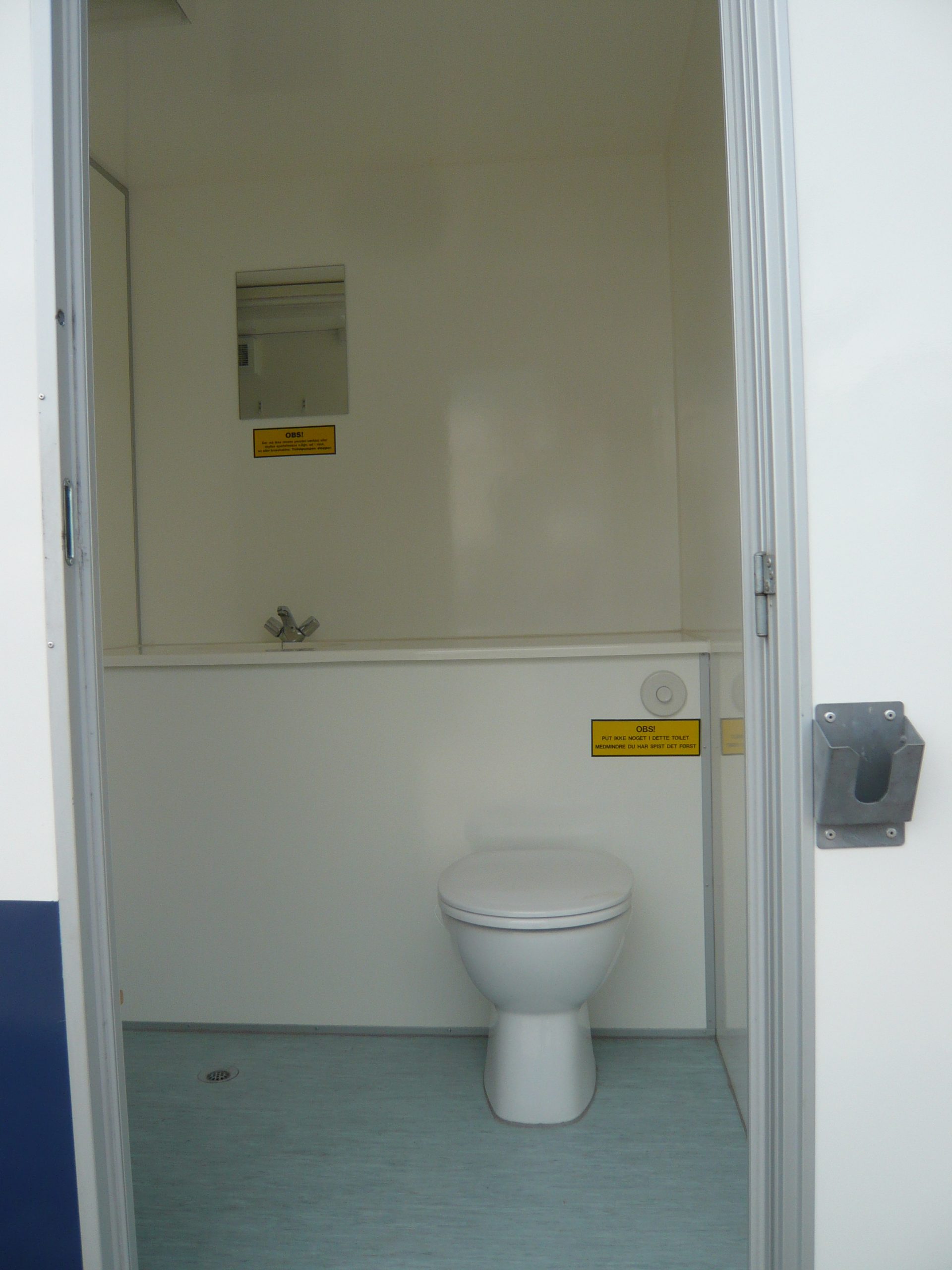 Innenansicht einer kleinen, weißen Toilette mit geschlossenem Deckel und mehreren gelben Hinweisschildern an der Wand