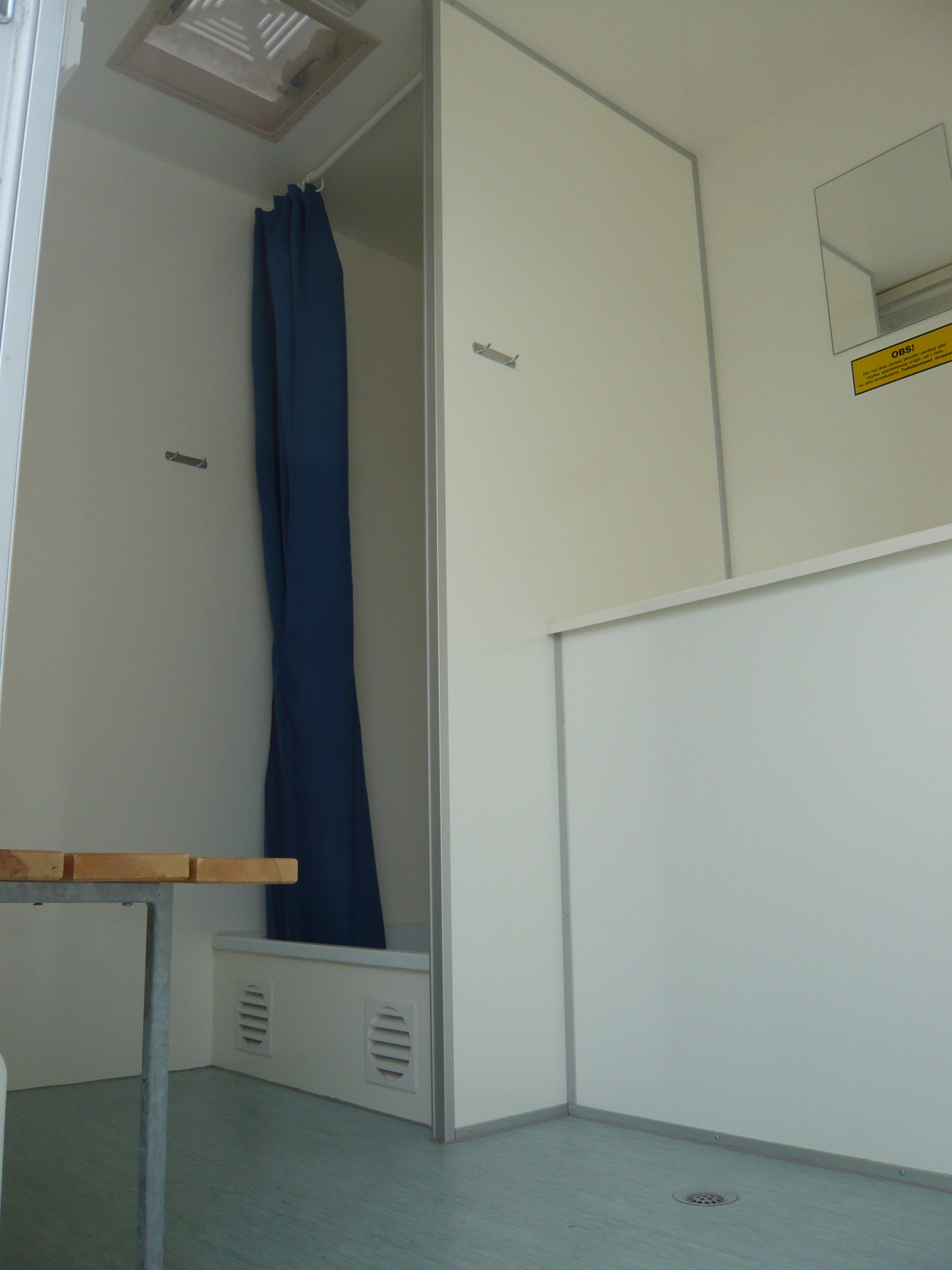 Blick in einen hellen Raum mit blauer Gardine, weißen Wänden, zwei Türen mit Schildern und einem Holztisch mit Metallgestell
