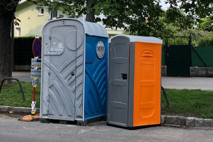 Zwei mobile Toiletten nebeneinander auf einem Gehweg, eine blau-graue mit Aufschrift 'MPA' und eine orange-graue