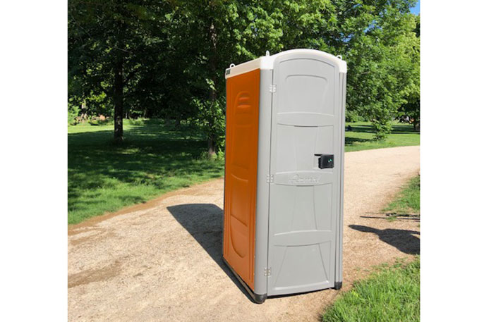 Freistehende mobile Toilette mit grauer Tür und orangefarbenen Seitenwänden auf einem Weg in einem Park mit Bäumen im Hintergrund