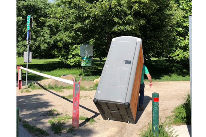 Person schiebt eine mobile Toilette über einen Weg mit Schranke in einem Park mit Bäumen im Hintergrund