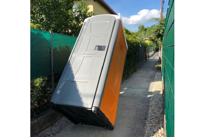 Eine umgefallene mobile Toilette steht schräg auf einem schmalen Gehweg zwischen einem grünen Zaun und einer Hecke.