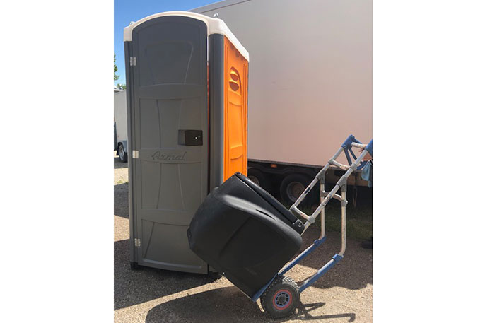 Mobile Toilette in Grau und Orange neben einem Sackkarren mit schwarzem Behälter auf Asphalt
