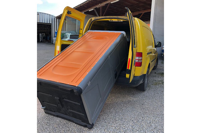 Gelber Lieferwagen mit geöffneter Heckklappe, aus dem eine große, orange-graue tragbare Toilette herausragt